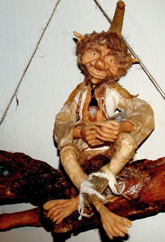 A small carved duende figure, a mischievous goblin from Spanish and Latin American folklore, seated cross-legged on a branch