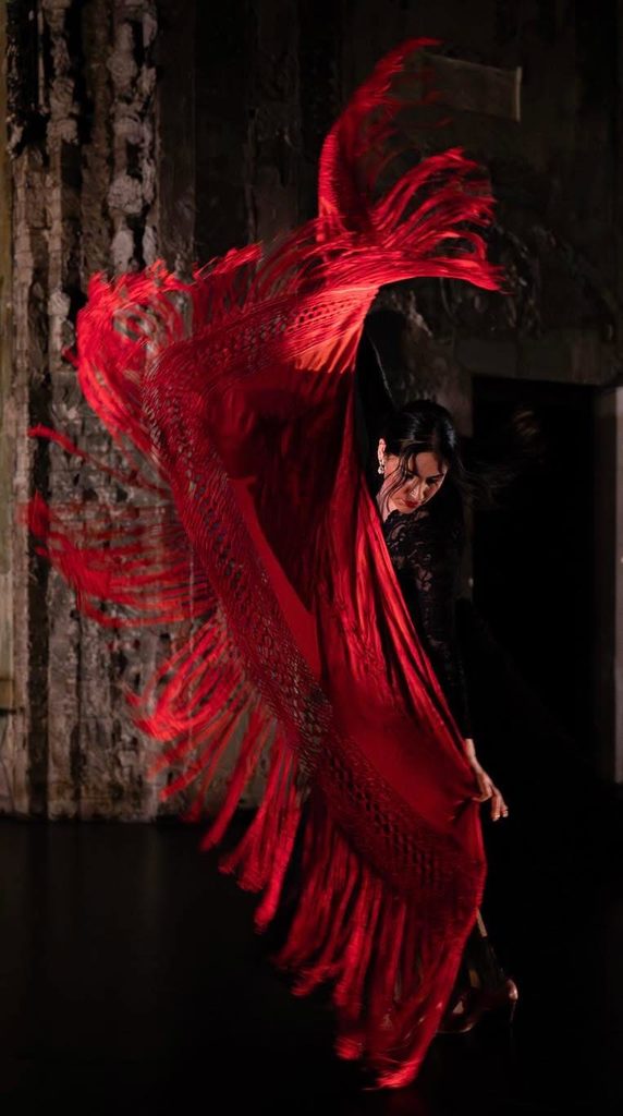A flamenco dancer in black spins a large red mantón, the fringed fabric fanned dramatically behind her on a dark stage