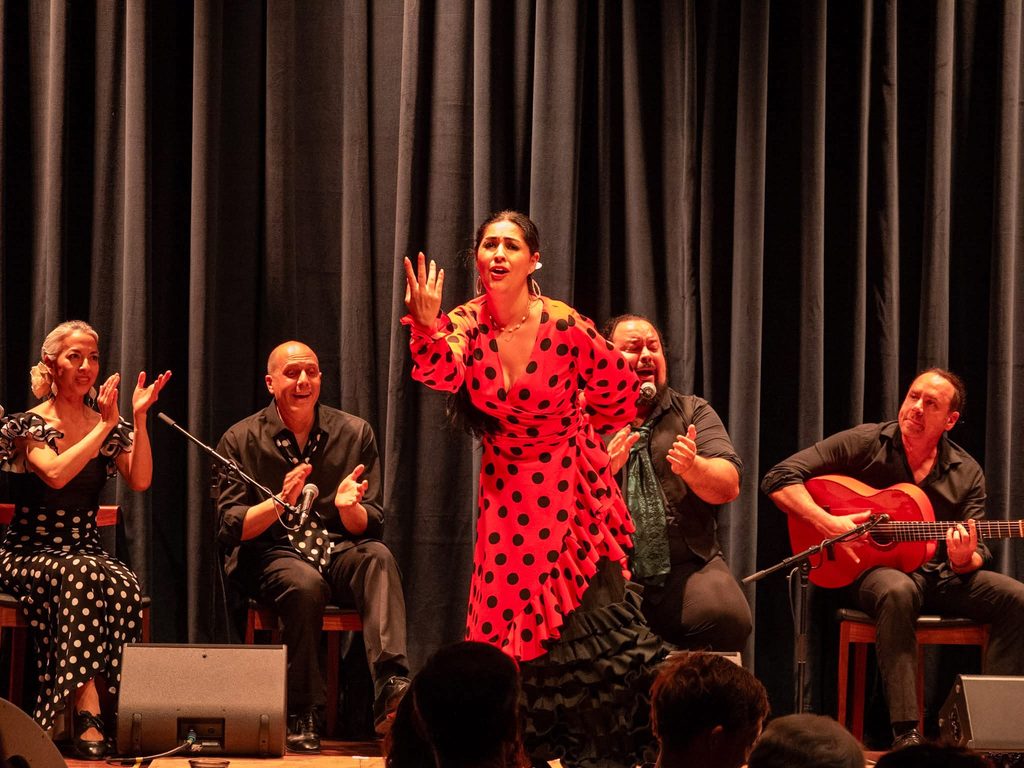 Flamenco X vocalist in a red polka-dot dress sings with raised hand, flanked by seated musicians clapping palmas and a guitarist